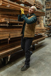 Woman wearing Dovetail Workwear Old School Trucker Vest in Vintage Brown over a teal hoodie, smiling while holding a gold award in a lumber warehouse