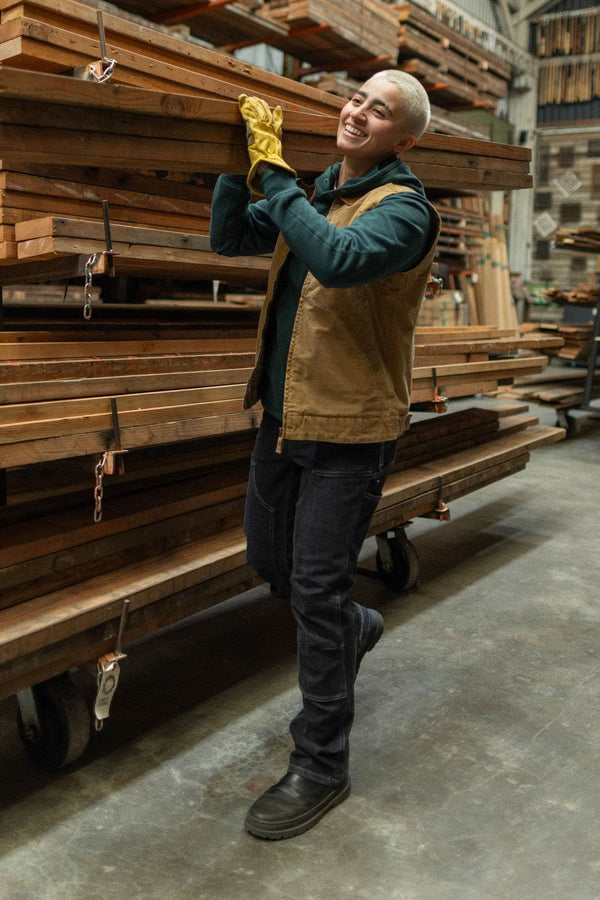 Woman wearing Dovetail Workwear Old School Trucker Vest in Vintage Brown over a teal hoodie, smiling while holding a gold award in a lumber warehouse