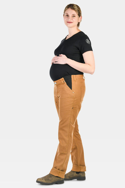 Side view of a pregnant woman wearing Dovetail Workwear's Anna Maternity Work Pants in Saddle Brown Canvas with a black t-shirt and work boots