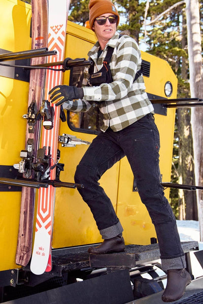Woman wearing Dovetail Workwear's Britt Utility Pant in Black Stretch Thermal Denim with reinforced knees, standing on a yellow ski lift carrying skis in a snowy mountain setting