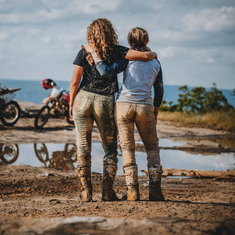 Two people standing on a dirt track with mud on their clothing, facing a puddle that reflects their and two off-road motorcycles in the background under a cloudy sky.