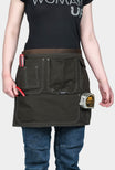 Dovetail Workwear Tool Apron in Kodiak Brown worn by a woman, displaying multiple pockets, hammer loops, and twine hole eyelets at the waist
