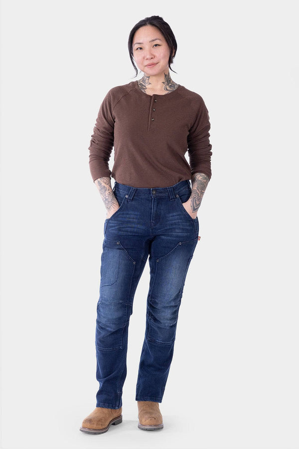 Woman wearing Dovetail Workwear Britt X Graphene Denim Pant in indigo with brown henley shirt and tan boots, standing against white background