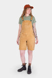 Tan Sandstone Utility Short Overalls by Dovetail Workwear with 9" inseam, worn by a woman with a green t-shirt, rust cap, and black boots against a white background