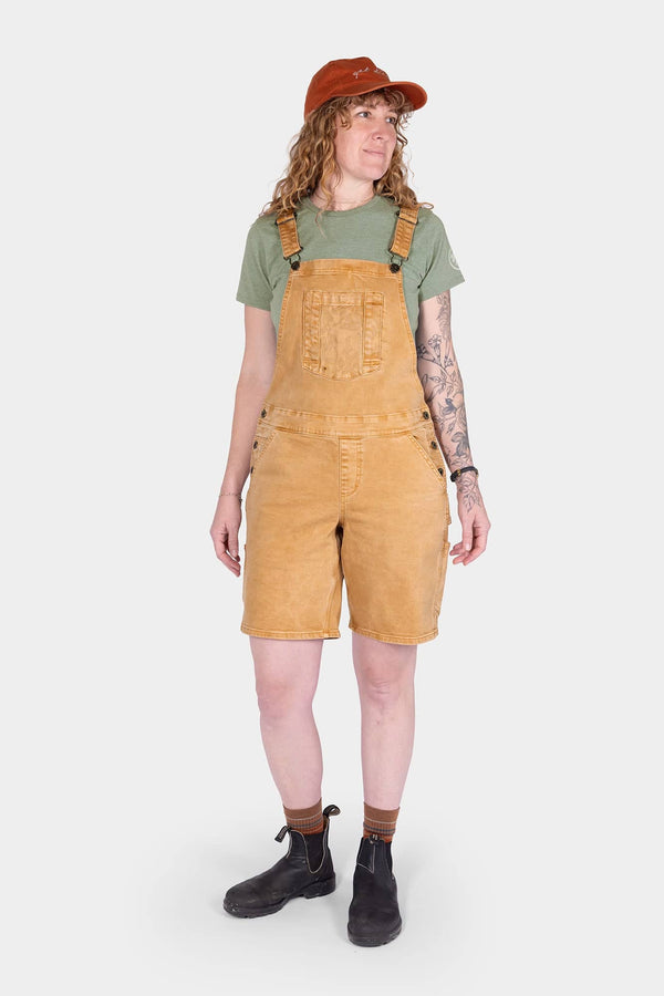 Tan Sandstone Utility Short Overalls by Dovetail Workwear with 9" inseam, worn by a woman with a green t-shirt, rust cap, and black boots against a white background