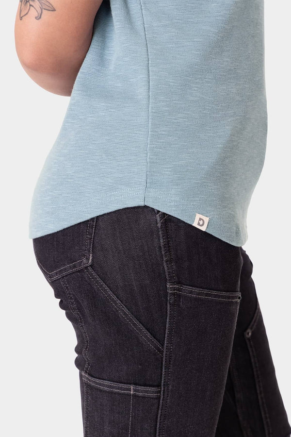 Side view of a woman wearing the Dovetail Workwear Old School Summer Henley Shirt in Shore Blue, paired with dark denim work pants