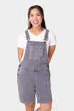 Woman smiling while wearing Dovetail Workwear Utility Short Overalls in Driftwood Grey with white t-shirt, featuring bib pocket, suspenders, and multiple functional pockets