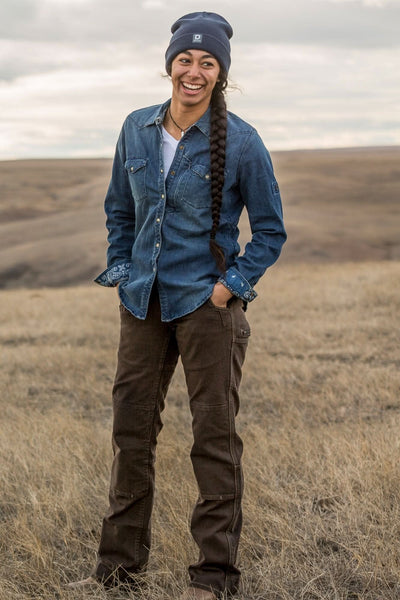 Woman wearing Dovetail Workwear Day Construct Pant in Brown Canvas with denim shirt and beanie, standing in an open field