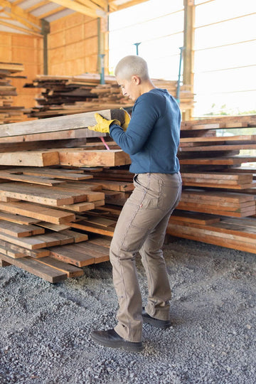 Woman wearing Dovetail Workwear's Honed Long Sleeve Thermal Henley in Atlantic Blue, inspecting lumber at a worksite, showcasing the waffle knit thermal base layer and reinforced elbows.