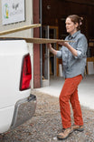 Woman wearing Dovetail Workwear Anna Taskpant in Paprika Orange, carrying lumber while loading a white van at a job site