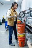 Woman in a workshop wearing the Vintage Brown Old School Trucker Vest by Dovetail Workwear, demonstrating the vest's deep hand pockets and light quilted insulation while operating machinery