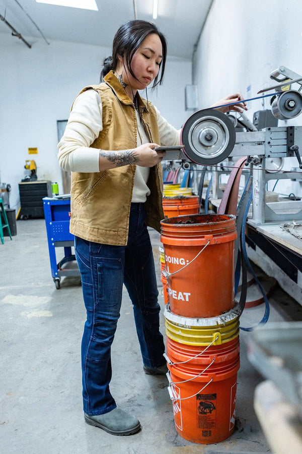 Woman in a workshop wearing the Vintage Brown Old School Trucker Vest by Dovetail Workwear, demonstrating the vest's deep hand pockets and light quilted insulation while operating machinery