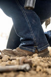 Woman wearing John Deere X Dovertail Workwear Into Town Bootcut Jeans in Classic Indigo Blue with brown boots standing in dirt, showing the bootcut fit and Free Hem™ split design