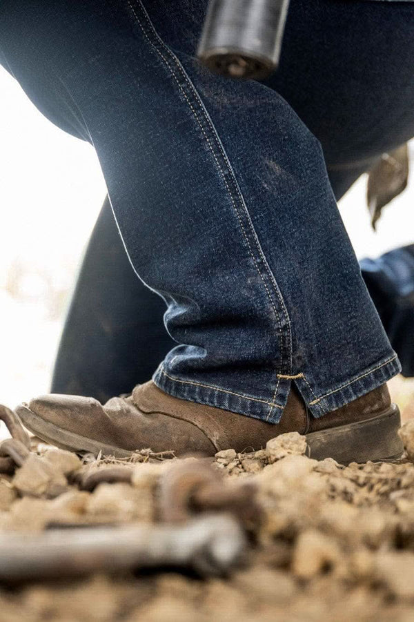 Woman wearing John Deere X Dovertail Workwear Into Town Bootcut Jeans in Classic Indigo Blue with brown boots standing in dirt, showing the bootcut fit and Free Hem™ split design