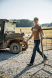 Woman wearing John Deere X Dovetail Workwear Into Town Bootcut Jeans in Indigo Denim, standing by a farm gate with a John Deere vehicle in the background