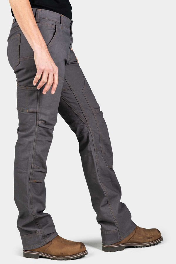 Side view of a woman wearing Dovetail Workwear's Britt Utility Pant in FR Grey Canvas, showcasing cargo-style pockets, straight leg fit, and reinforced knees with brown work boots