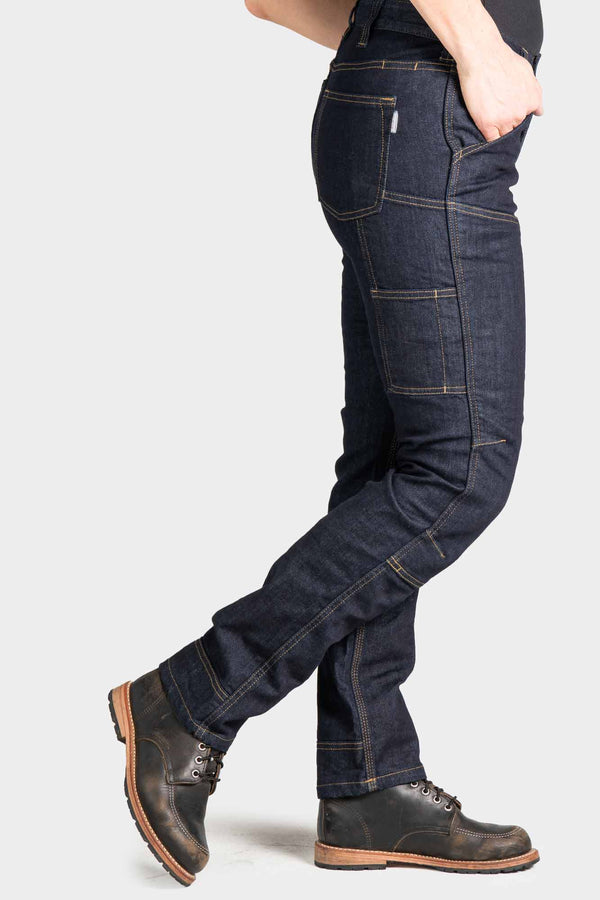 Side view of a woman wearing Dovetail Workwear's Britt Utility Pant in FR Denim Indigo, showing cargo pockets, reinforced knees, and straight leg silhouette with work boots
