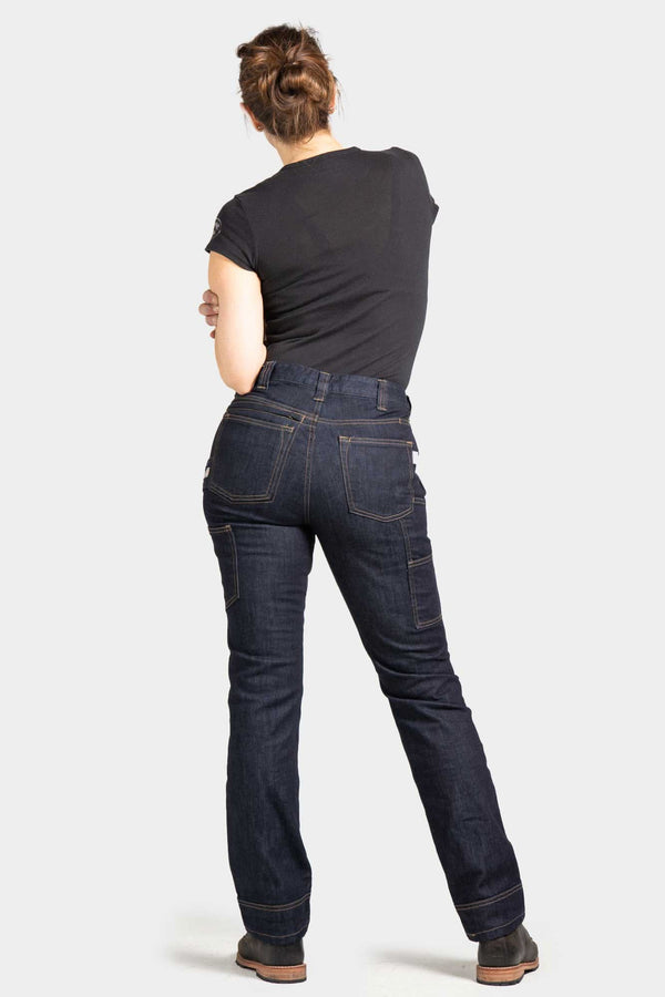 Back view of a woman wearing Dovetail Workwear's Britt Utility Pant in Indigo FR Denim, showing the straight leg fit, reinforced knees, and cargo-style pockets.